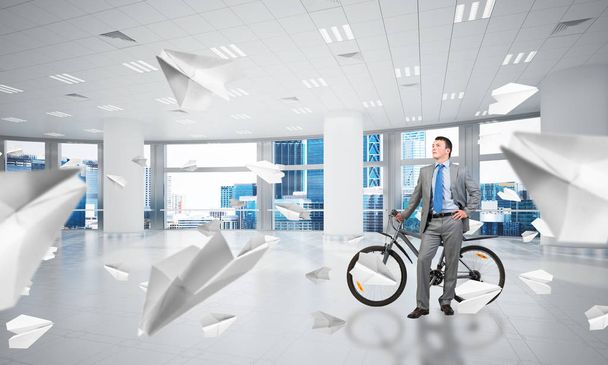 Young man wearing business suit and tie with bike standing at comfortable coworking space with flying paper planes. Happy businessman with bicycle at modern office interior with big windows. - Foto, imagen