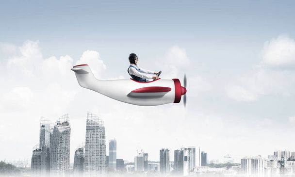 Επιχειρηματίας που κάθεται στην καμπίνα του μικρού αεροπλάνου και κρατά το τιμόνι. Αστείος άνθρωπος με καπέλο πιλότου και προστατευτικά γυαλιά οδήγηση αεροπλάνο έλικα πάνω από την πόλη. Σύγχρονη μητρόπολη με ψηλά κτήρια και πύργους - Φωτογραφία, εικόνα