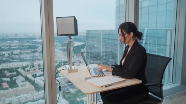 business woman works in a modern office located on a high floor of a skyscraper. attractive girl working on a laptop while sitting at the table by a large window. - Materiał filmowy, wideo