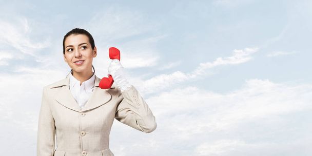 Attractive young woman holding red retro phone. Call center operator in white business suit posing with telephone on skyscape background. Hotline telemarketing. Business assistance and consultation. - Valokuva, kuva