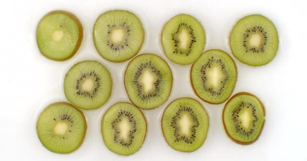 A água espirra em câmara lenta. Vista superior: vários círculos de kiwi é lavado com água em um fundo branco. frutos picados
 - Filmagem, Vídeo