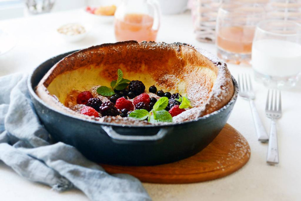 Fresh homemade dutch baby pancake with powdered sugar, fresh berries and mint for breakfast with a glass of milk and apple juice on a light background. Delicious  breakfast in a sunny interior. - 写真・画像