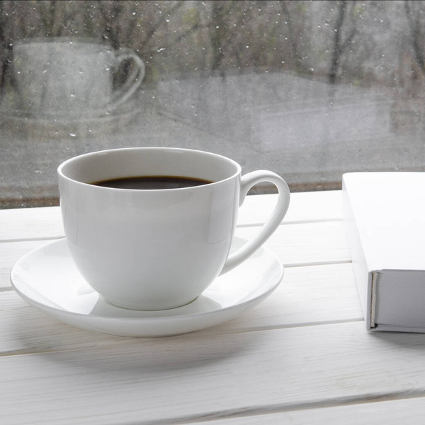 Gezellig thuis stilleven: witte kop hete koffie en wit geopende boek over vensterbank tegen sneeuw landschap buiten. - Foto, afbeelding