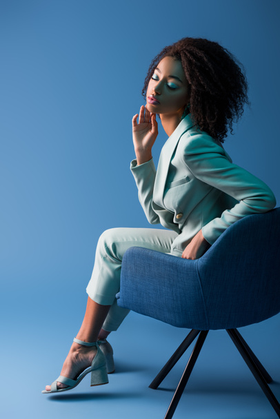 african american woman sitting on armchair with closed eyes on blue background  - Фото, зображення