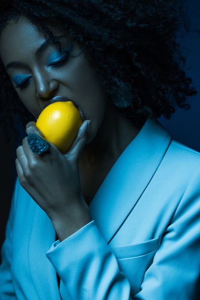 attractive african american woman with makeup eating lemon isolated on blue - Фото, изображение