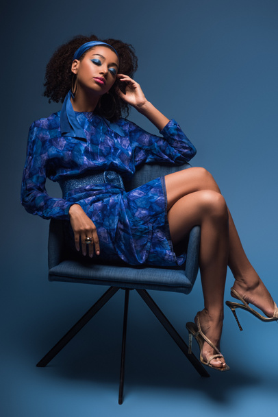 ελκυστική Αφροαμερικανή γυναίκα με κλειστά μάτια κάθεται στην πολυθρόνα σε μπλε φόντο  - Φωτογραφία, εικόνα