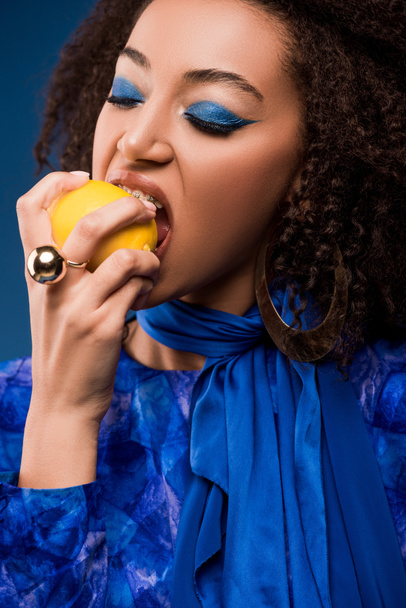 Αφροαμερικανή γυναίκα με μακιγιάζ τρώει νόστιμο λεμόνι που απομονώνεται στο μπλε - Φωτογραφία, εικόνα