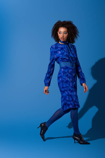 attractive african american woman in dress walking on blue background  - Zdjęcie, obraz