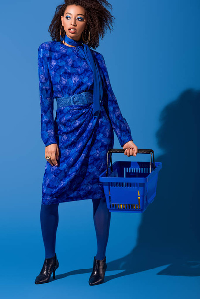 african american woman holding shopping basket on blue background  - Foto, Imagen
