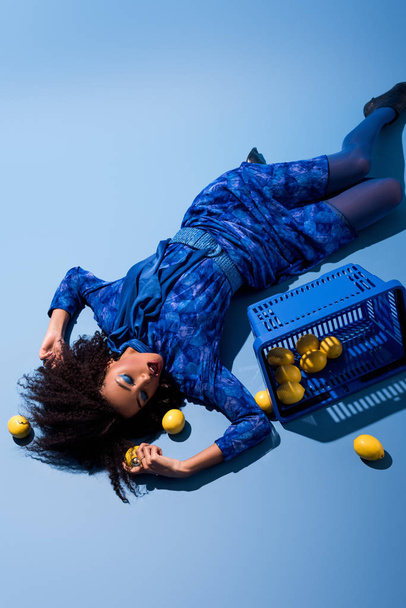hoge hoek uitzicht van Afrikaans amerikaanse vrouw liggen met winkelmandje en citroenen op blauwe achtergrond  - Foto, afbeelding