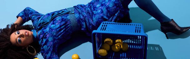 panoramic shot of african american woman lying with shopping basket and lemons on blue background  - Photo, Image