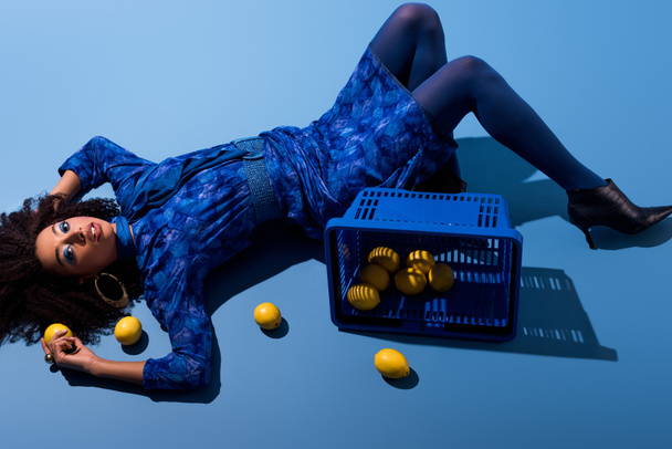 african american woman lying with shopping basket and lemons on blue background  - 写真・画像