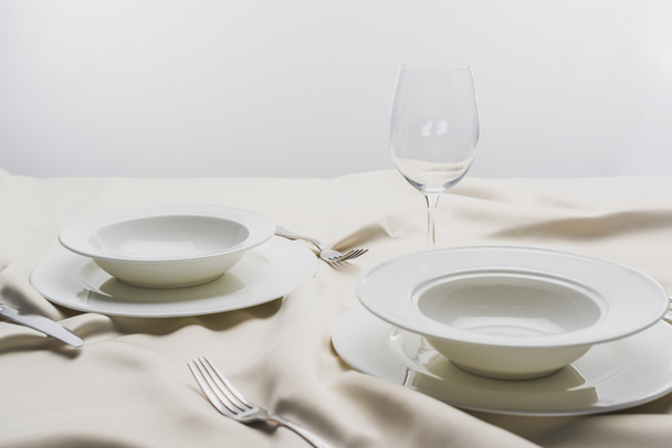 Plates with cutlery and clear wine glass on white tablecloth on grey background - 写真・画像