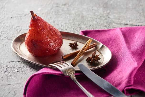 delicious pear in wine with cinnamon and anise on silver plate on grey concrete surface with pink napkin, knife and fork - Photo, image
