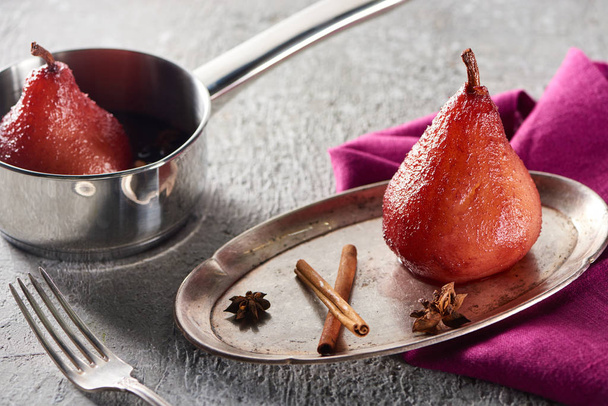delicious pear in wine with cinnamon and anise on silver plate and in stewpot on grey concrete surface with pink napkin and fork - Photo, image