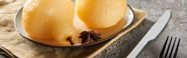 delicious pear in wine with anise and cinnamon served on plate on napkin with knife and fork on grey concrete surface, panoramic shot - Zdjęcie, obraz
