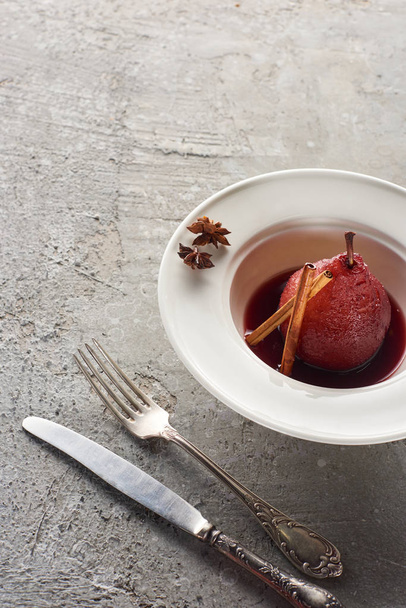 νοστιμότατο αχλάδι σε κόκκινο κρασί με κανέλα και γλυκάνισο σε πιάτο σε γκρι τσιμεντένια επιφάνεια με μαχαιροπήρουνα - Φωτογραφία, εικόνα