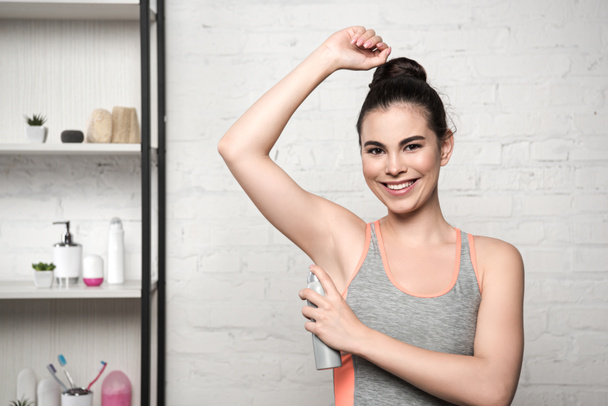 mujer alegre sonriendo a la cámara mientras aplica desodorante en la axila
 - Foto, Imagen