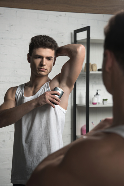 joven con camisa blanca sin mangas aplicando desodorante en la axila mientras mira el espejo
 - Foto, Imagen