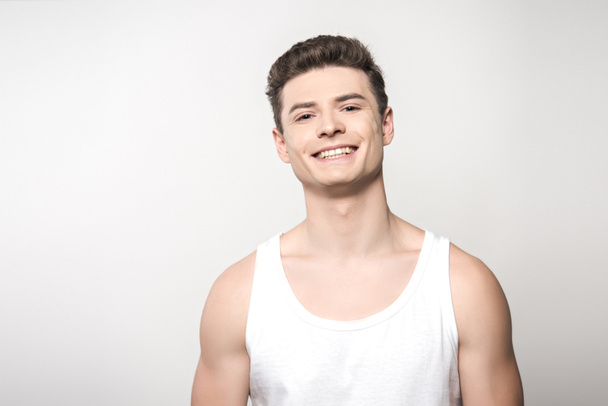 handsome man in white sleeveless shirt smiling at camera isolated on grey - Foto, immagini