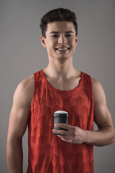 young man in red sleeveless shirt holding deodorant while smiling at camera isolated on grey - Fotó, kép