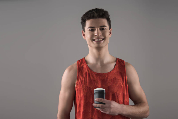 joven con camisa roja sin mangas sosteniendo desodorante mientras sonríe a la cámara aislada en gris
 - Foto, imagen