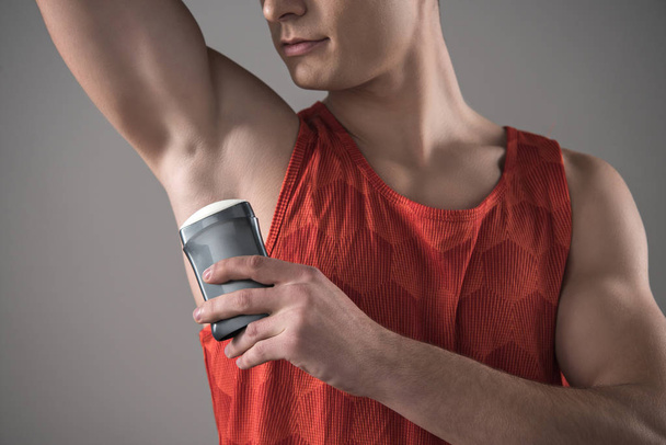 cropped view of young man in red sleeveless shirt applying deodorant on underarm isolated on grey - Foto, Bild