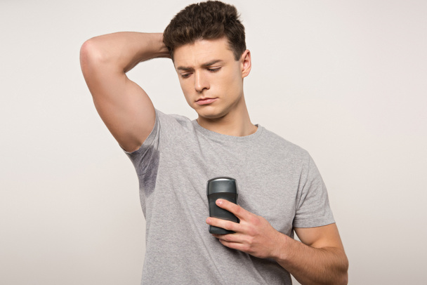displeased man in grey t-shirt with sweaty underarm holding deodorant isolated on grey - Photo, Image
