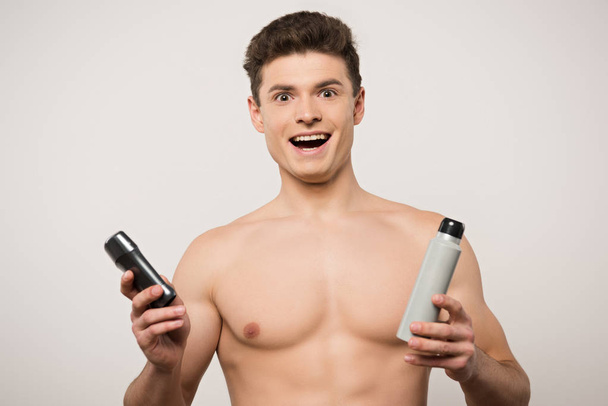 podekscytowany mężczyzna bez koszulki patrząc w kamerę trzymając dezodoranty odizolowane na szaro - Zdjęcie, obraz