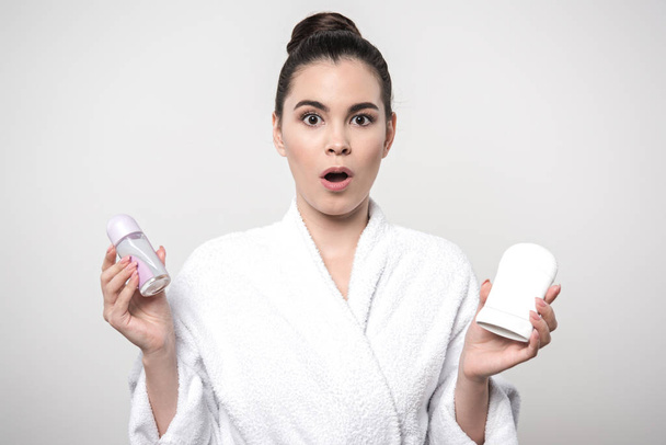 surprised woman in bathrobe holding deodorants while looking at camera isolated on grey - Foto, imagen