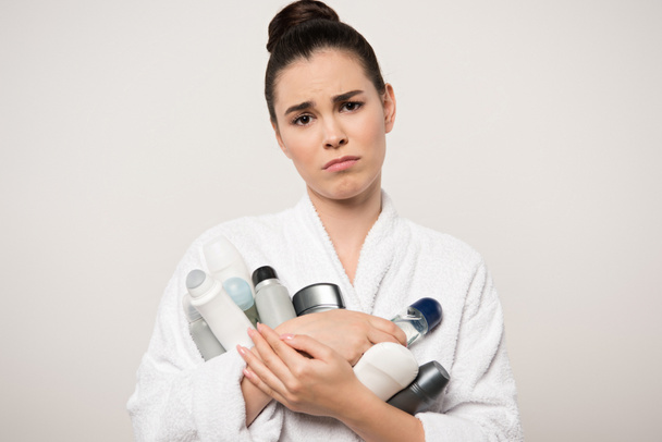 mujer disgustada en albornoz sosteniendo diferentes desodorantes mientras mira a la cámara aislada en gris
 - Foto, imagen