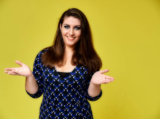 Portrait of a pretty brunette woman with a beautiful hairstyle and with excellent makeup in a dark blue blouse on a yellow background. It stands showing hands with a smile in front of the camera. - Foto, afbeelding