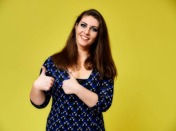Portrait of a pretty brunette woman with a beautiful hairstyle and with excellent makeup in a dark blue blouse on a yellow background. It stands showing hands with a smile in front of the camera. - Foto, immagini