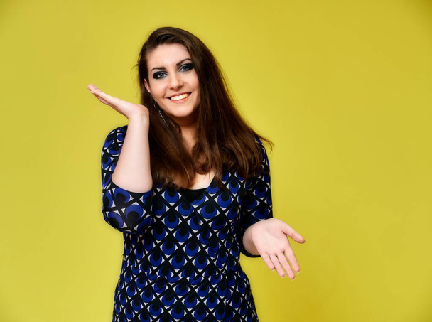 Portrait of a pretty brunette woman with a beautiful hairstyle and with excellent makeup in a dark blue blouse on a yellow background. It stands showing hands with a smile in front of the camera. - Photo, Image