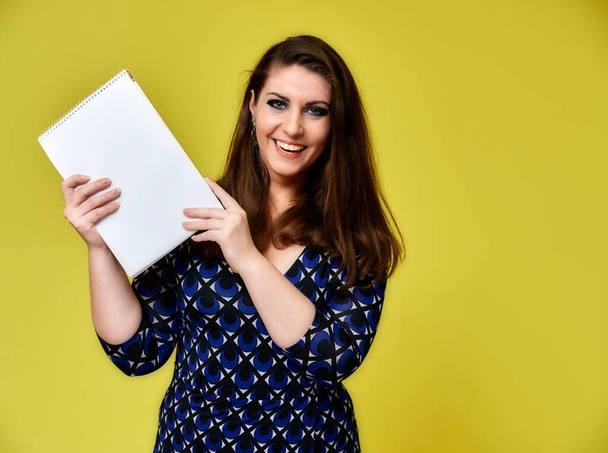 Portrait of a pretty brunette woman with a folder in her hands with a beautiful hairstyle and with excellent makeup in a dark blue blouse on a yellow background. Stands with a smile. - Photo, Image