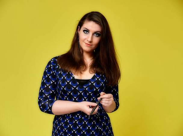 Portrait of a pretty brunette woman with a beautiful hairstyle and with excellent makeup in a dark blue blouse on a yellow background. He stands with a smile and shows with his hands. - Foto, imagen