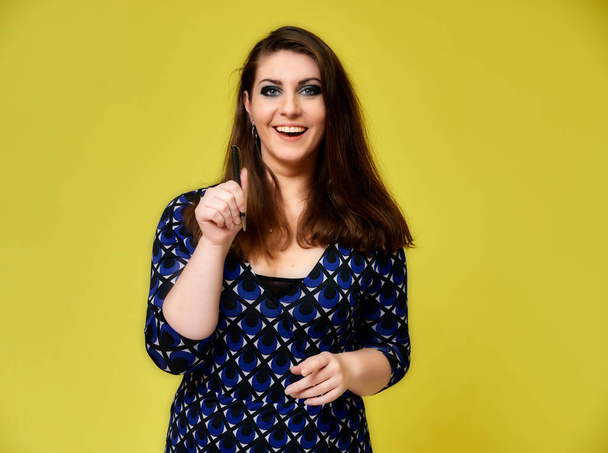 Portrait of a pretty brunette woman with a beautiful hairstyle and with excellent makeup in a dark blue blouse on a yellow background. He stands with a smile and shows with his hands. - Foto, afbeelding