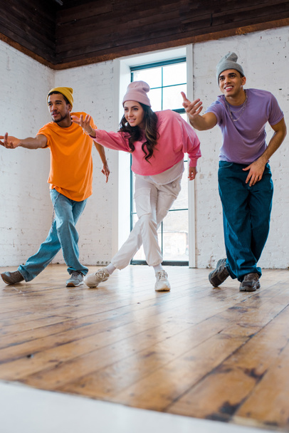 stylowa dziewczyna gestykulując podczas breakdance ze szczęśliwymi wielokulturowymi mężczyznami w czapkach  - Zdjęcie, obraz