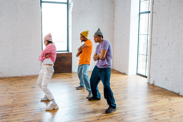 стильна дівчина з схрещеними руками танцює з красивими мультикультурними чоловіками в капелюхах
  - Фото, зображення