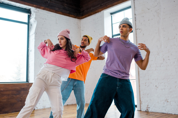 girl breakdancing and gesturing with stylish multicultural men in hats  - Foto, Bild