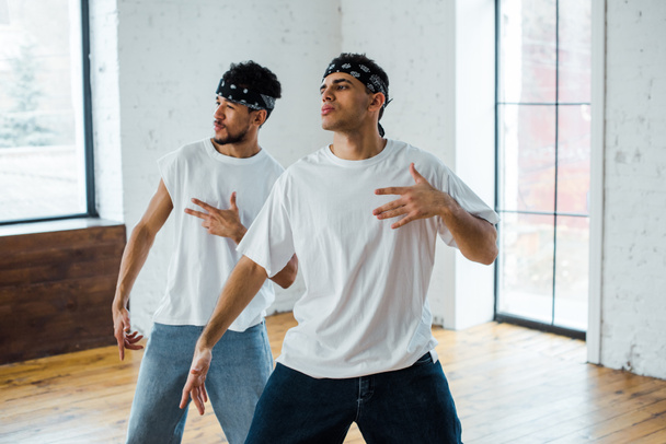 knappe multiculturele dansers in hoofdbanden gebaren en poseren tijdens het dansen hip-hop - Foto, afbeelding