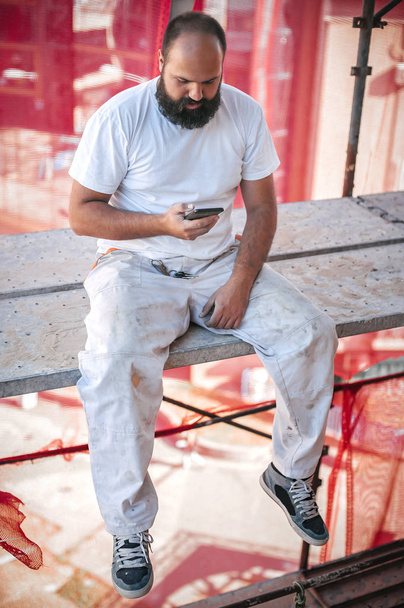 Building site worker sitting high on the scaffolding, resting and using mobile phone - Φωτογραφία, εικόνα