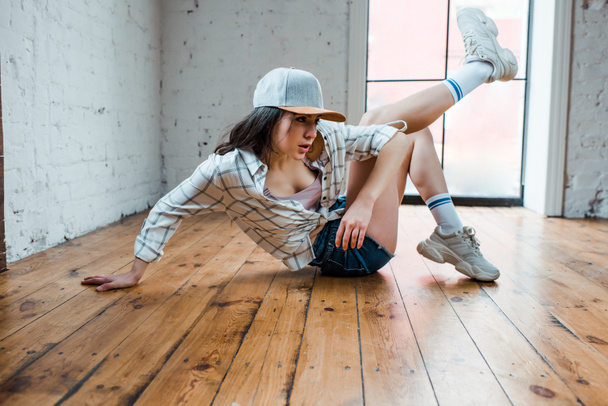 beautiful dancer in cap sitting on floor and dancing hip-hop in dance studio  - Fotó, kép