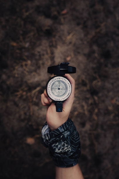 Hand of man with a scarf, holding older metal compass with arrow pointing west. Background is out of focus with coniferous forest environment. - Photo, image