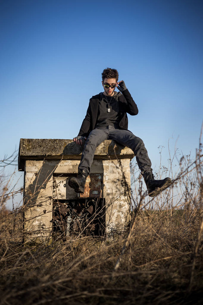 zerzauster, charismatischer junger Mann in schwarzem Outfit, mit schurkischem Gesichtsausdruck, sitzt auf einem verlassenen Betonklotz mit reinblauem Himmel im Hintergrund und verdorrter Vegetation ringsum. - Foto, Bild