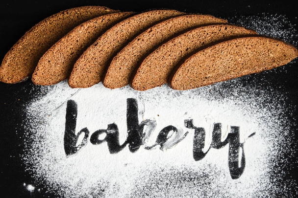 Inscripción panadería sobre harina de trigo blanco dispersa Pan de centeno en rodajas sobre fondo oscuro
 - Foto, imagen
