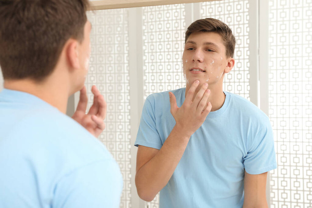 Teen guy with acne problem applying cream near mirror indoors - Foto, Imagen