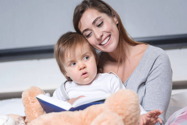 Young beautiful mother reads to baby the book before going to sleep in bed - Foto, Bild