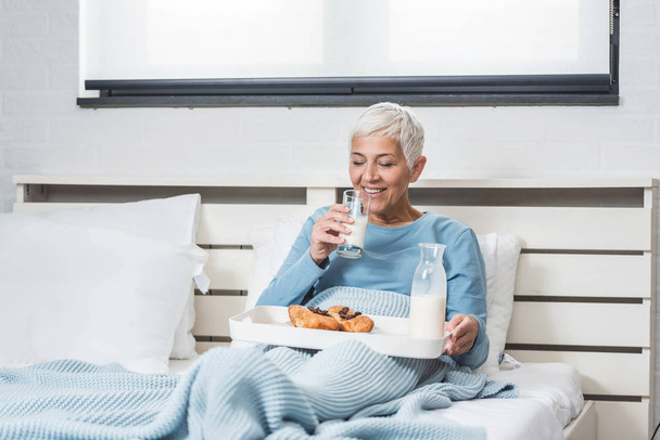 Senior woman eating breakfast in bed holding plate with croissant and drink glass of milk - Foto, imagen