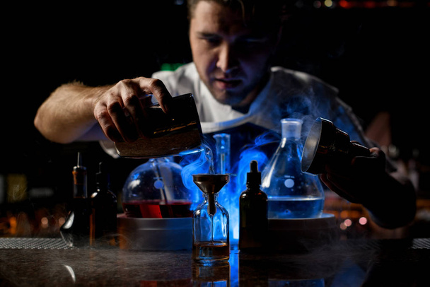 Bartender pouring a transparent alcoholic drink from the steel shaker to the little bottle under blue light - Foto, Imagen
